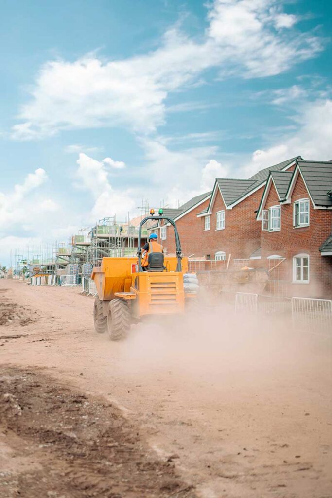 A yellow construction vehicle drives along a dusty path near newly built brick houses and scaffolding under a blue sky.