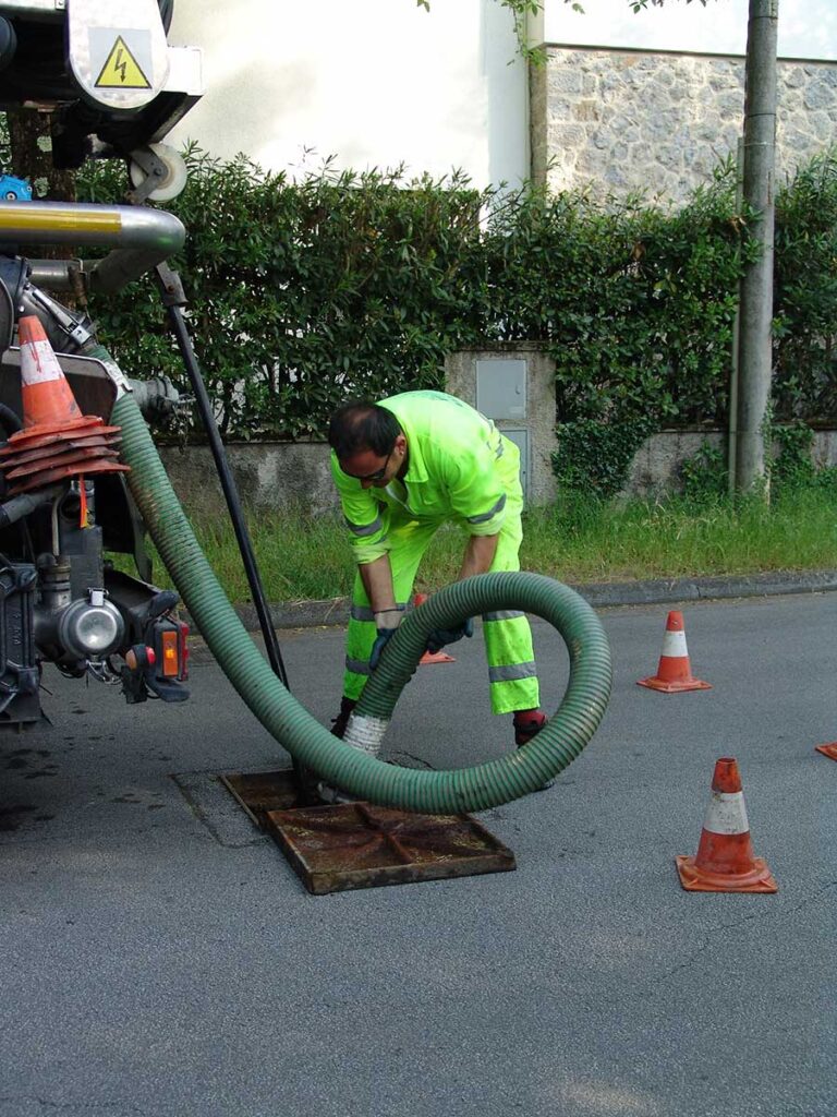 Worker in a fluorescent suit handling a hose near a manhole, with traffic cones and machinery nearby.